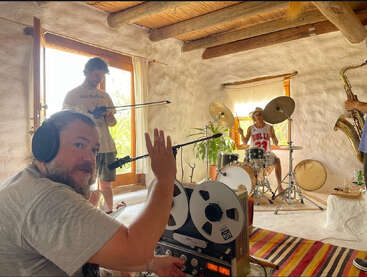 Quatro músicos ensaiam em uma sala aconchegante e iluminada pelo sol. Um deles grava com equipamento antigo, enquanto os outros tocam bateria, violino e saxofone. A atmosfera é criativa e descontraída.