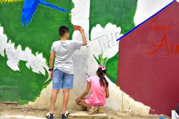 Deux enfants sont en train de peindre une fresque colorée sur un mur. La peinture murale représente un perroquet bleu, des fleurs blanches et des mots en rouge. Les enfants ont l'air concentrés.