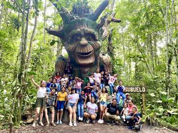 Un groupe de personnes, principalement des enfants, se réunit joyeusement dans une forêt luxuriante à côté d'une sculpture d'arbre géante au visage amical. Un panneau indique "Abuelo".