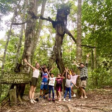 Un groupe d'enfants souriants et deux adultes posent joyeusement dans une forêt luxuriante à côté d'un panneau en bois indiquant "Bosque de Huayo" et d'une sculpture d'arbre.