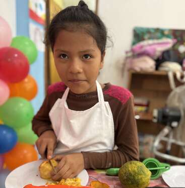 Une jeune fille portant un tablier blanc coupe des fruits sur une table. Des ballons colorés se trouvent à l'arrière-plan, créant une atmosphère festive et joyeuse.