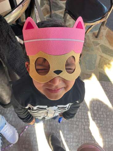 A young child wearing a pink and tan cat mask smiles with eyes closed, dressed in a skeleton shirt, standing indoors with chairs and sunlight nearby.