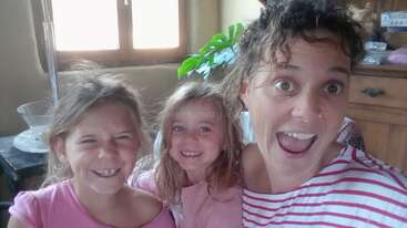 Uma mulher sorridente e duas meninas posam para uma selfie alegre em um ambiente interno. A luz quente do sol brilha através da janela atrás delas, criando uma atmosfera aconchegante e alegre.