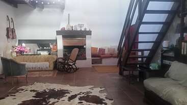 Esta sala de estar aconchegante tem lareira, cadeira de balanço, sofás, violões na parede, tapete de couro, estante, escadas e itens decorativos, criando uma atmosfera acolhedora.