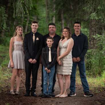 Seis personas posan juntas al aire libre en un camino forestal, vestidas de manera formal e informal. El grupo sonríe, rodeado de frondosos árboles verdes y una suave luz natural.