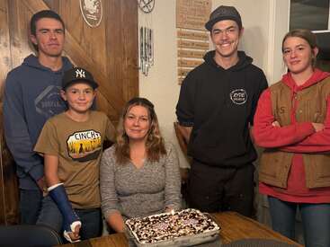 Eine Frau sitzt an einem Tisch mit einer verzierten Torte, umgeben von vier lächelnden Kindern oder Jugendlichen. Es scheint sich um ein Familienfest oder einen Geburtstag zu handeln.