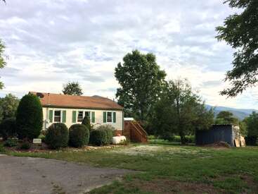 Ein kleines Haus mit hellbraunen Fassaden und einem braunen Dach steht inmitten von grünen Büschen und Bäumen, während im Hintergrund die Berge unter einem teilweise bewölkten Himmel zu sehen sind.