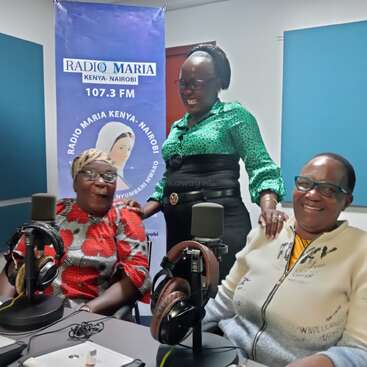 Three women are cheerful in a radio studio, seated around microphones. A blue “Radio Maria Kenya-Nairobi 107.3 FM” banner is visible in the background.