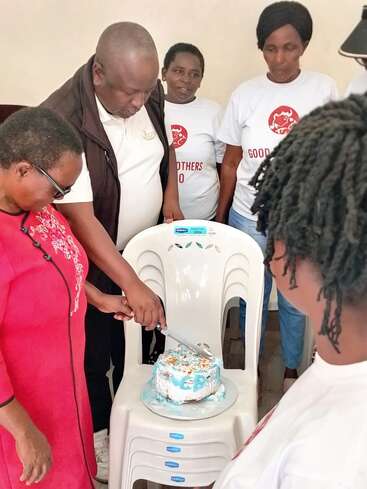 A group of people gather while a woman in a red dress cuts a cake placed on a white chair, creating a warm celebratory atmosphere.