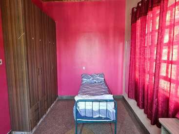 This image shows a simple bedroom with pink walls, a single metal bed, purple bedding, large wardrobe, maroon curtains, and sunlight streaming through the window.
