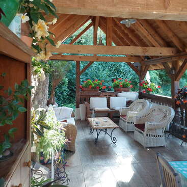 Un acogedor patio cubierto de estilo rústico cuenta con muebles de mimbre, asientos acolchados, plantas de un verde intenso y flores de vivos colores, lo que crea un espacio exterior relajado y acogedor.