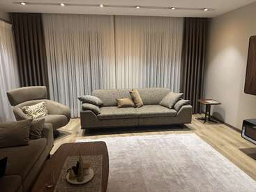 Uma sala de estar moderna com um sofá cinza, poltrona combinando, tapete creme, mesa de centro, mesa lateral, cortinas neutras e iluminação suave, criando uma atmosfera aconchegante.