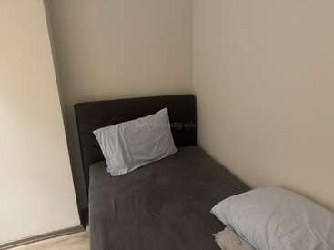 Esta imagem mostra uma cama pequena, bem arrumada, com cabeceira escura e roupa de cama cinza, colocada no canto de um quarto simples, com paredes creme.