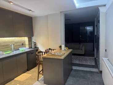 Cozinha moderna com armários marrons, bancadas de madeira e ilha. O layout aberto leva você à sala de estar com um sofá bege. Iluminação sutil, decoração minimalista, organizada e convidativa.