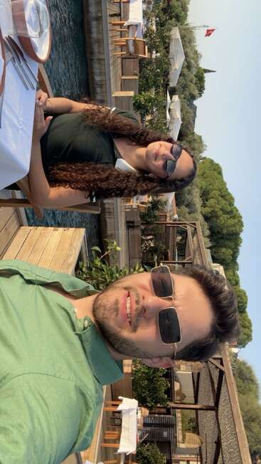 Un hombre y una mujer se sientan junto al agua en un restaurante al aire libre, sonrientes y con gafas de sol. La vegetación y las mesas de madera crean un ambiente relajado y soleado.
