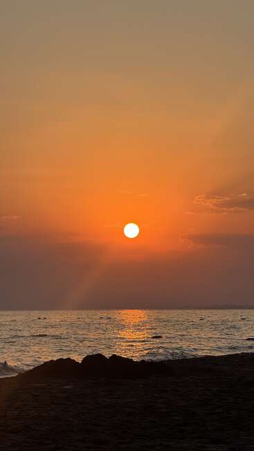 A glowing orange sunset over a calm ocean. The sun reflects on the water, fluffy clouds in the sky, and a dark sandy beach foreground. Peaceful scenery.