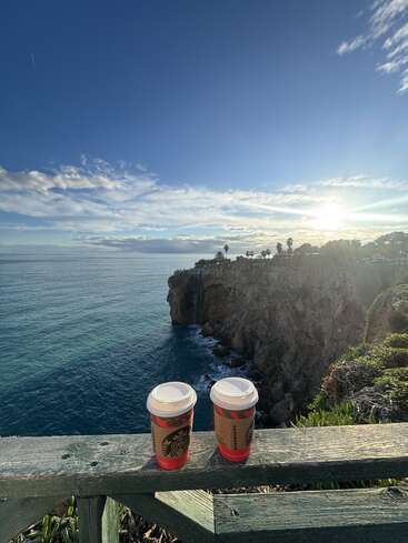 Two Starbucks coffee cups rest on a wooden railing overlooking a dramatic ocean cliff, with bright sunlight, blue sky, scattered clouds, and lush coastal greenery.