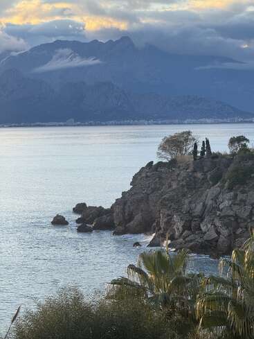 A rocky coastline with palm trees overlooks calm blue water. Distant mountains rise under dramatic cloudy skies, with a city faintly visible across the bay.