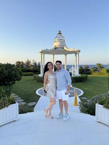 A smiling couple poses together in front of a white gazebo surrounded by lush greenery, with the ocean and a clear blue sky in the background.