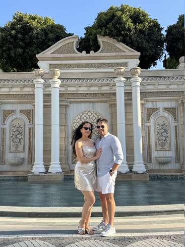 A couple stands smiling in front of a beautiful, ornate fountain with classical pillars and lush trees, dressed stylishly in summer outfits under a clear blue sky.