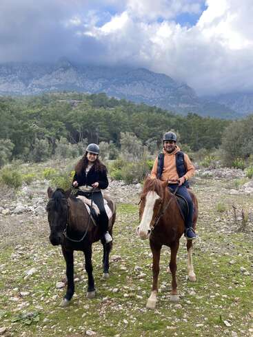 A man and a woman ride horses through a rocky, green landscape. Both wear helmets. Behind them, dense forest and cloudy mountains set a dramatic scene.
