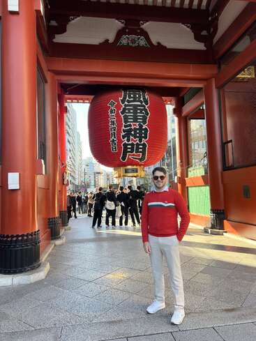A man in a red sweater poses beneath a giant red lantern at Kaminarimon Gate, Senso-ji Temple, Tokyo. Crowds and city buildings are visible behind.
