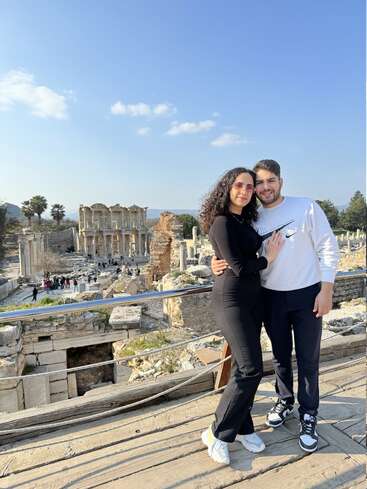 A couple poses affectionately in front of ancient ruins under a clear blue sky. The historical site, with columns and crowds, creates a stunning, romantic backdrop.