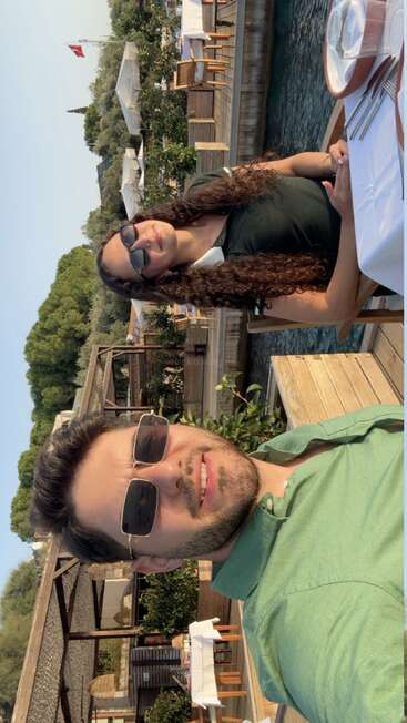 A man and woman sit by the water at an outdoor restaurant, smiling and wearing sunglasses. Greenery and wooden tables create a relaxed, sunny atmosphere.