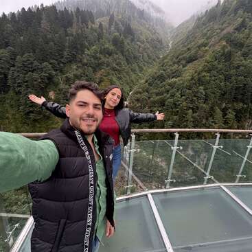 Una pareja sonriente se toma una selfie en un mirador acristalado con vistas a unas montañas verdes, frondosas y neblinosas y a un denso bosque, disfrutando juntos del paisaje natural.