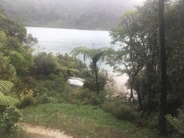 Un petit bateau blanc repose sur le rivage, entouré d'arbres et de fougères verdoyants. L'eau brumeuse et les collines forment un arrière-plan naturel et tranquille.