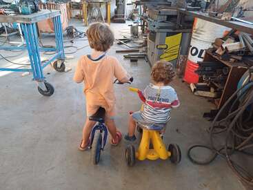 Two young children ride small bikes inside a cluttered workshop. They are seen from behind, surrounded by tools, cables, machinery, and metal work materials. Playful atmosphere.