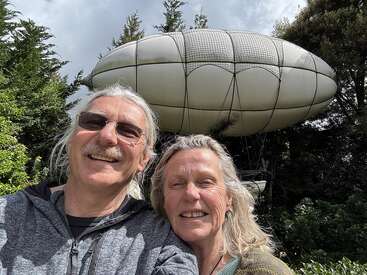 A smiling couple takes a selfie outdoors. Behind them is a unique, large, blimp-like structure among green trees, under a cloudy sky, giving an adventurous vibe.