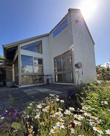 A modern white house with large windows sits under a bright blue sky. Wildflowers bloom in the foreground. Patio features a small table and chairs.