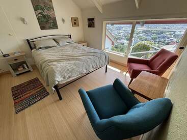 A cozy bedroom with a large bed, colorful rug, two comfy chairs, wooden floor, and large window offering a beautiful view of a cityscape outside.
