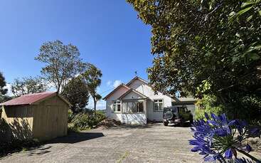 A white house with a front porch sits under a bright blue sky, surrounded by trees and greenery. A shed and parked car are visible.