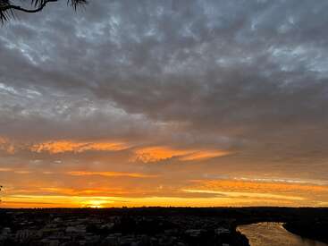 The image shows a breathtaking sunset, with golden and orange hues illuminating dramatic clouds over a river, a bridge, and a silhouetted cityscape below. Peaceful atmosphere.