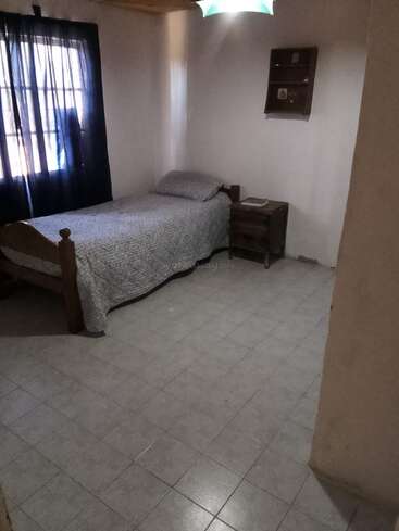 A small, simple bedroom with a single bed, patterned gray bedding, wooden nightstand, wall shelf, tiled floor, and dark curtain covering a sunlit window.