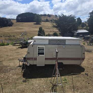 Uma caravana antiga está estacionada em um terreno seco e gramado, com colinas, árvores e galpões ao fundo, sob um céu parcialmente nublado, criando uma cena rural tranquila.