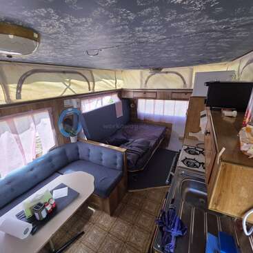 Essa imagem mostra o interior aconchegante de uma van de camping com uma pequena cozinha compacta, fogão, mesa de jantar, assentos almofadados, sofá-cama e luz entrando pelas cortinas.