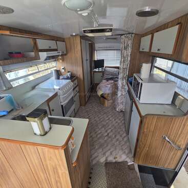 Esta imagen muestra el interior de una caravana clásica con paneles de madera, una pequeña cocina, una cocina, un microondas, un fregadero, armarios y una zona para dormir en la parte trasera.