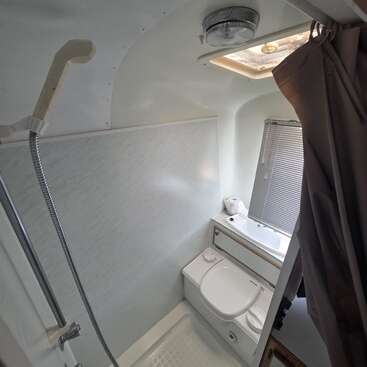En esta imagen se ve un cuarto de baño compacto de una autocaravana con un cabezal de ducha, un inodoro, un lavabo pequeño, persianas, un conducto de ventilación en el techo y una cortina de ducha marrón.