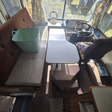 Esta imagen muestra la parte delantera del interior de una autocaravana o un autobús de estilo retro. Hay un sofá, una mesa, el asiento del conductor, una cubeta de plástico y cortinas estampadas. La luz del sol entra a raudales.