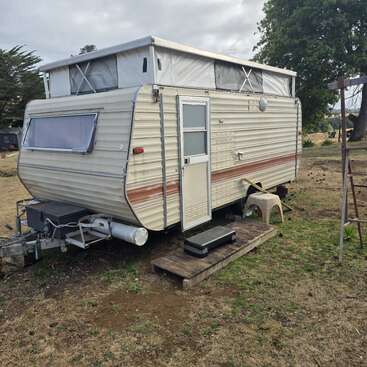 Una caravana vintage de color beige descansa sobre un trozo de césped. Tiene rejillas de ventilación en el techo, un pequeño porche de madera, unos escalones y un taburete de plástico cerca. Está rodeada de árboles.