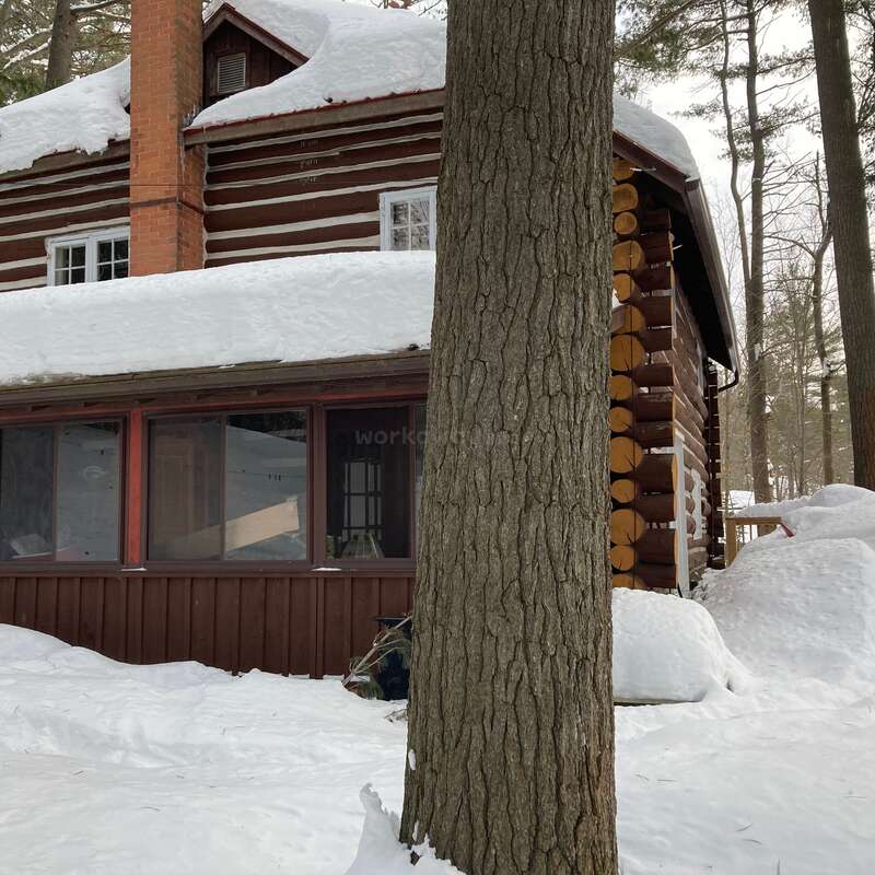 A cozy log cabin with a brick chimney is surrounded by deep snow and tall trees. Snow covers the roof and ground, giving a peaceful winter atmosphere.