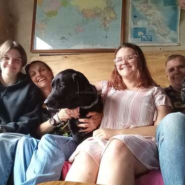 L'image représente une famille de quatre personnes, dont deux adultes et deux adolescents, assis sur un canapé avec deux chiens noirs, dans une atmosphère chaleureuse et confortable.