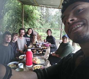 Das Bild zeigt eine Gruppe von sechs Personen, die sich um einen Tisch versammelt haben und gemeinsam eine Mahlzeit in einer ruhigen Umgebung im Freien genießen, umgeben von üppigem Grün.