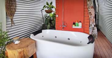 Cette image montre une baignoire moderne avec des appuie-tête noirs, entourée de murs en tôle ondulée, de plantes d'intérieur, d'un tabouret en bois, d'un mur orange, d'une douche et de produits de bain.