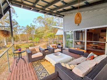 Une terrasse extérieure confortable équipée de meubles en osier avec coussins, d'une table, de tapis à motifs et offrant une vue sur la nature, abritée par un toit métallique et décorée d'une lampe suspendue.