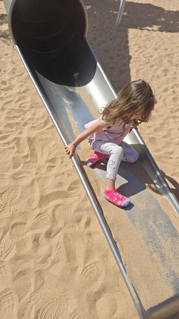 A young girl with long hair slides down a shiny metal slide onto sandy ground, wearing pink socks, white leggings with hearts, and a light pink shirt.