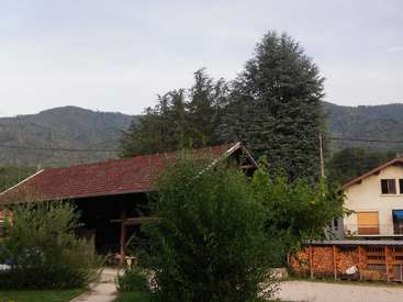 A imagem retrata uma cena rural serena com uma casa, um celeiro e um cenário montanhoso, com árvores e arbustos exuberantes que contribuem para o ambiente natural.